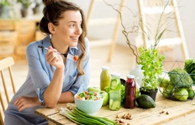 Mulher sorrindo à mesa com salada e sucos naturais, simbolizando escolhas saudáveis e consciência alimentar.