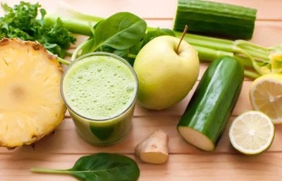 Bebida detox verde com abacaxi, maçã, pepino, limão e folhas frescas sobre mesa de madeira.