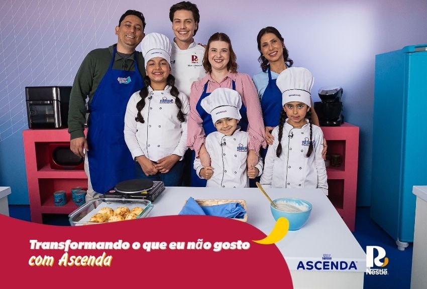 Adultos e crianças com uniformes de chef posam com receita em mesa, campanha “Transformando o que eu não gosto com Ascenda”.