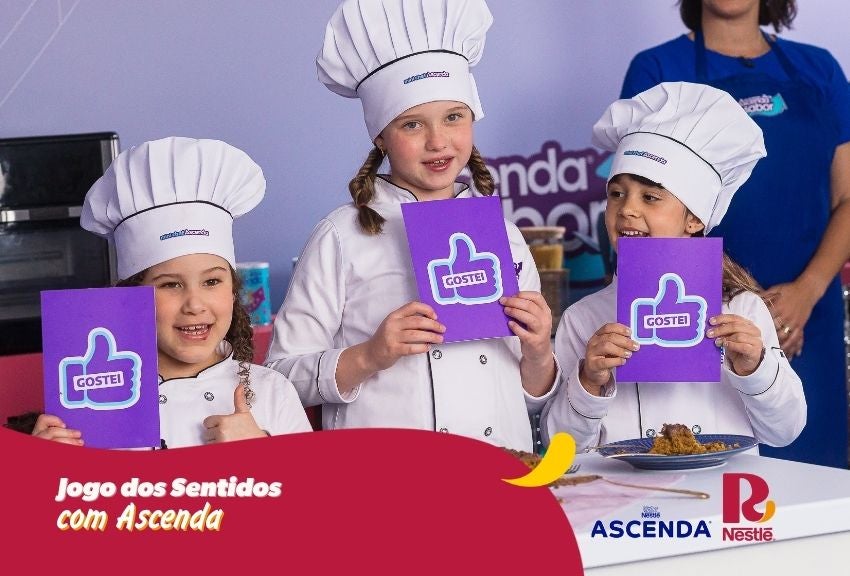 Três crianças com uniformes de chef seguram cards “Gostei” em atividade “Jogo dos Sentidos com Ascenda”.	