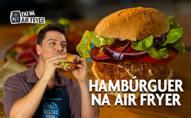 Receitas na Air Fryer Hambúrguer Hambúrguer preparado na Air Fryer com alface, tomate e cebola-roxa, com pessoa segurando o lanche ao lado.