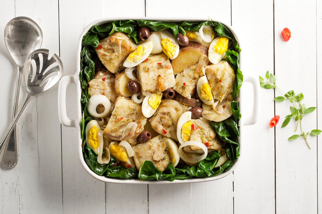 Imagem de uma travessa quadrada branca com bacalhau, couve, azeitonas e ovos sobre uma mesa de madeira branca.