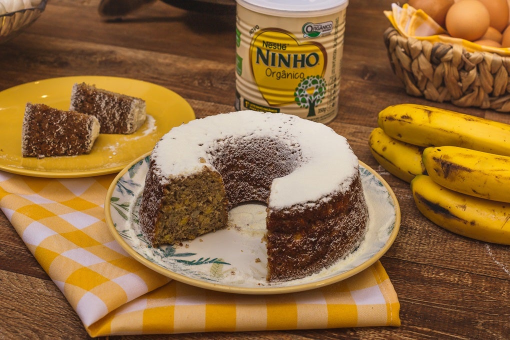 Imagem de um bolo de banana sobre um prato, com bananas ao lado e uma lata de Leite Ninho.