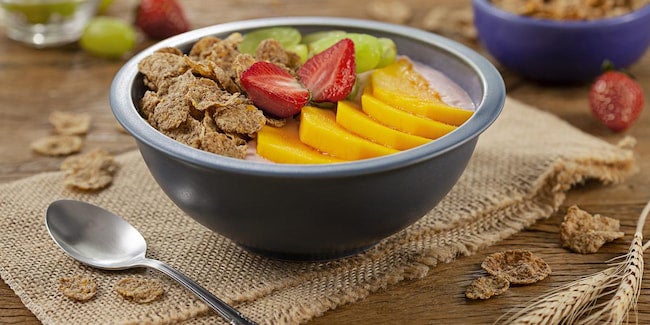 Bowl azul com iogurte, cereal e frutas como manga, morango e uva-verde.