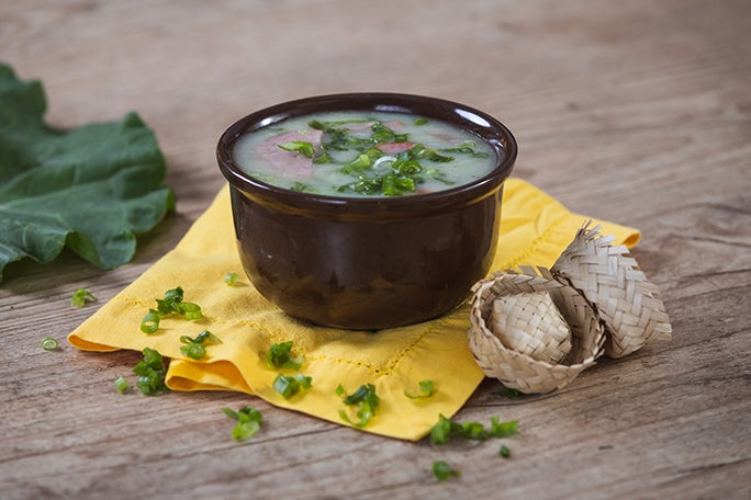 Caldo verde servido em cumbuca marrom sobre guardanapo amarelo, decorado com cheiro-verde e mini chapéus de palha ao lado.