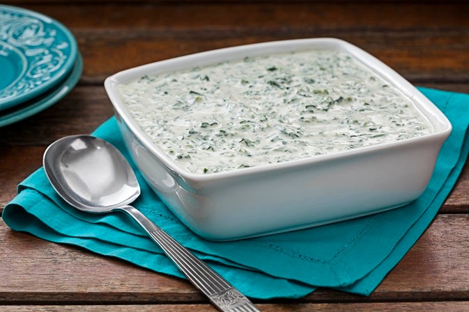 Imagem de uma travessa quadrada branca com creme de espinafre e uma colher ao lado.