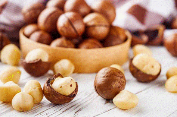 Bowl com macadâmias com casca e descascadas em superfície de madeira clara.