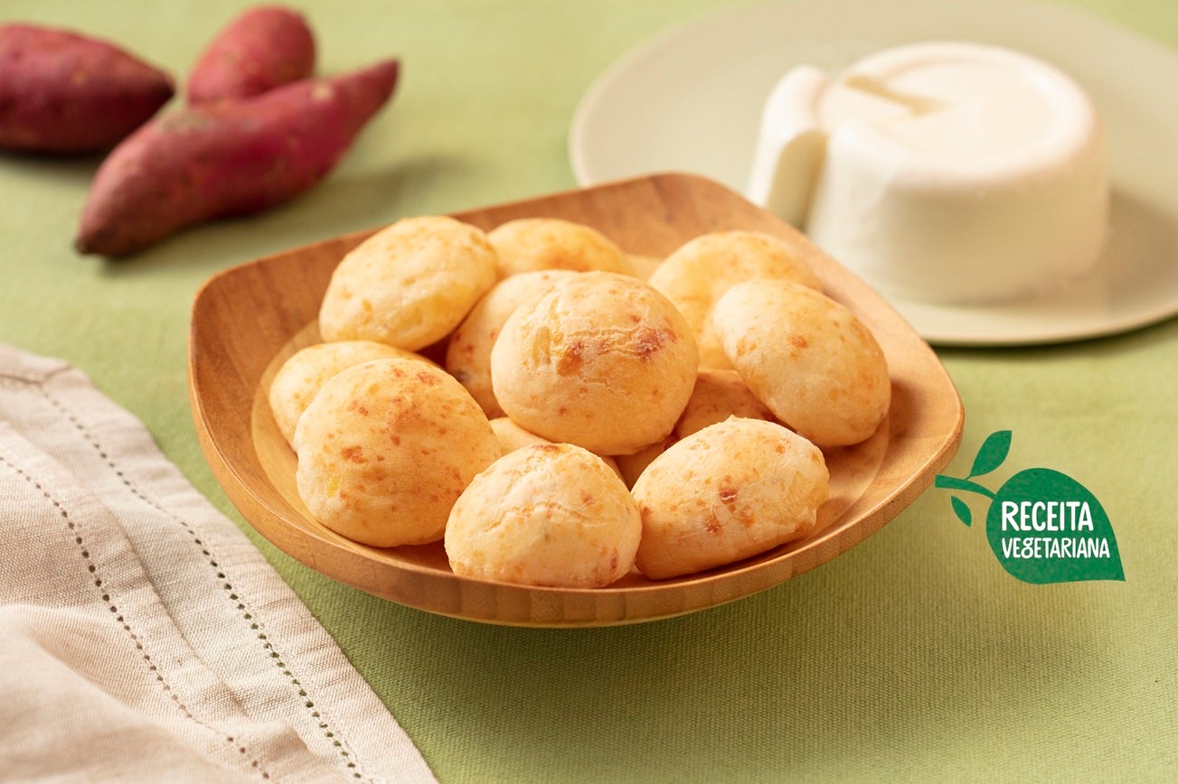 Imagem de um bowl de madeira com pães de queijo, batatas-doces ao fundo e um prato de queijo fresco.