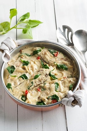 Imagem de uma panela com pescada ao molho cremoso de curry e espinafre sobre uma tábua de madeira branca.