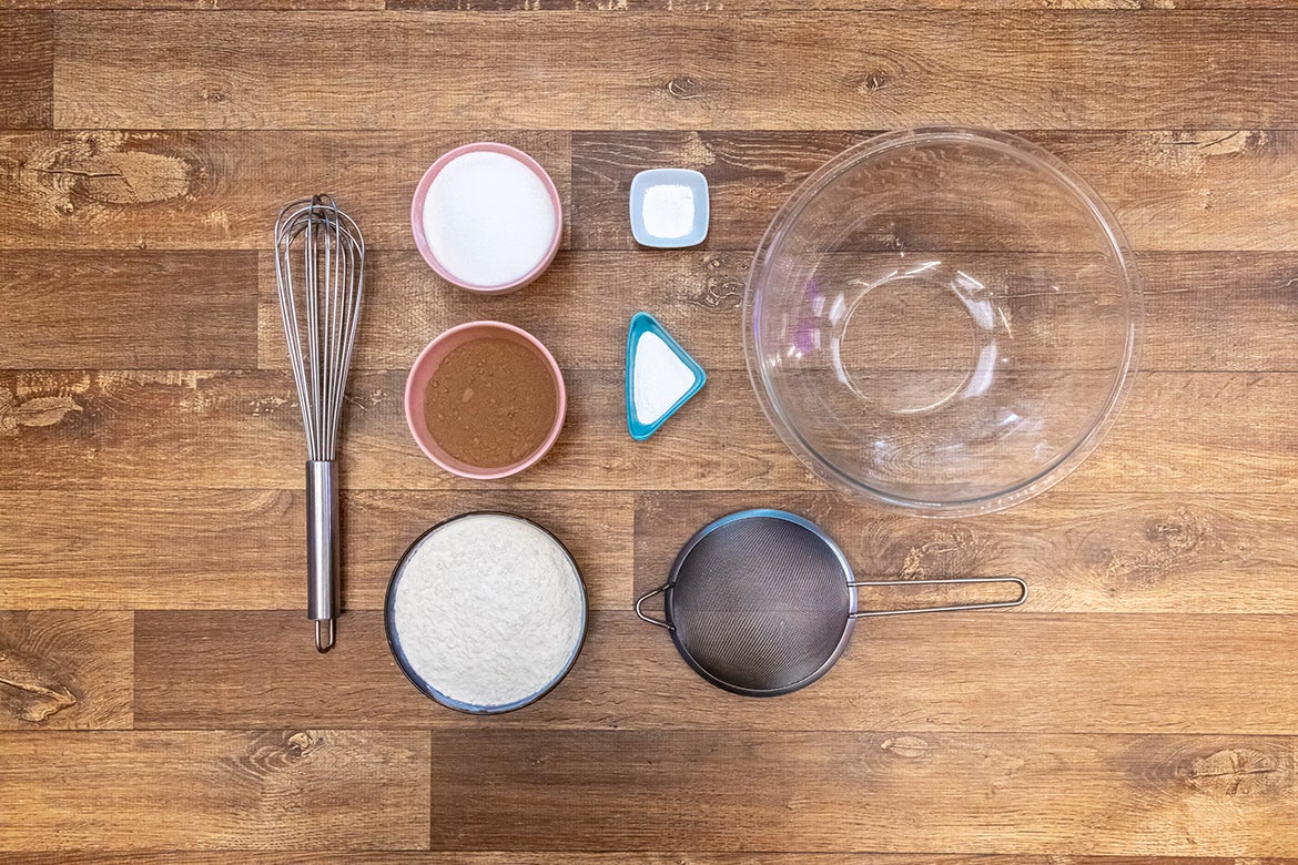 Foto vista de cima de todos os ingredientes secos dispostos em uma bancada de madeira ao lado de um recipiente grande de vidro transparente e de um batedor de arame grande