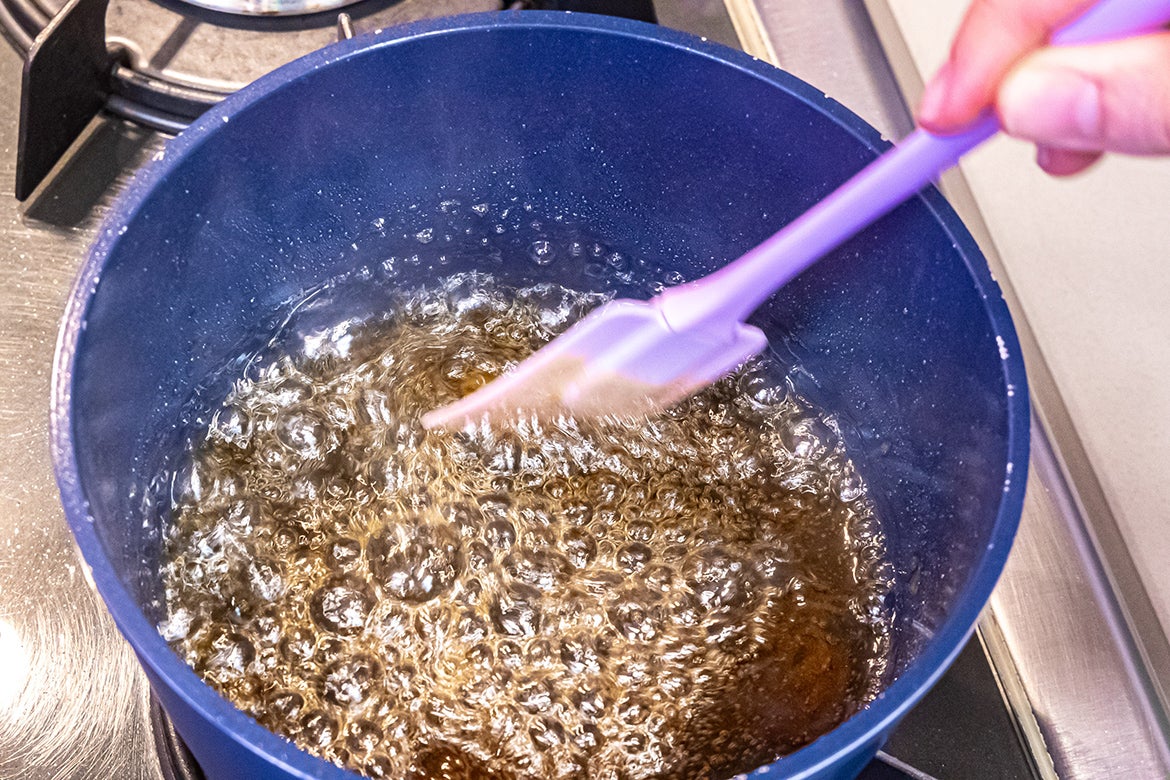 Foto aproximada da calda de caramelo fervendo em uma panela azul sendo mexida por uma espátula de silicone lilás