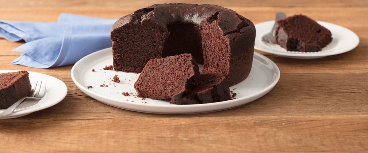 Fotografia em tons de marrom e branco de uma bancada de madeira clara com um paninho azul claro e um prato branco redondo com o bolo de chocolate. Aos lados dois pratos brancos pequenos com fatias de bolo de chocolate e um garfo.