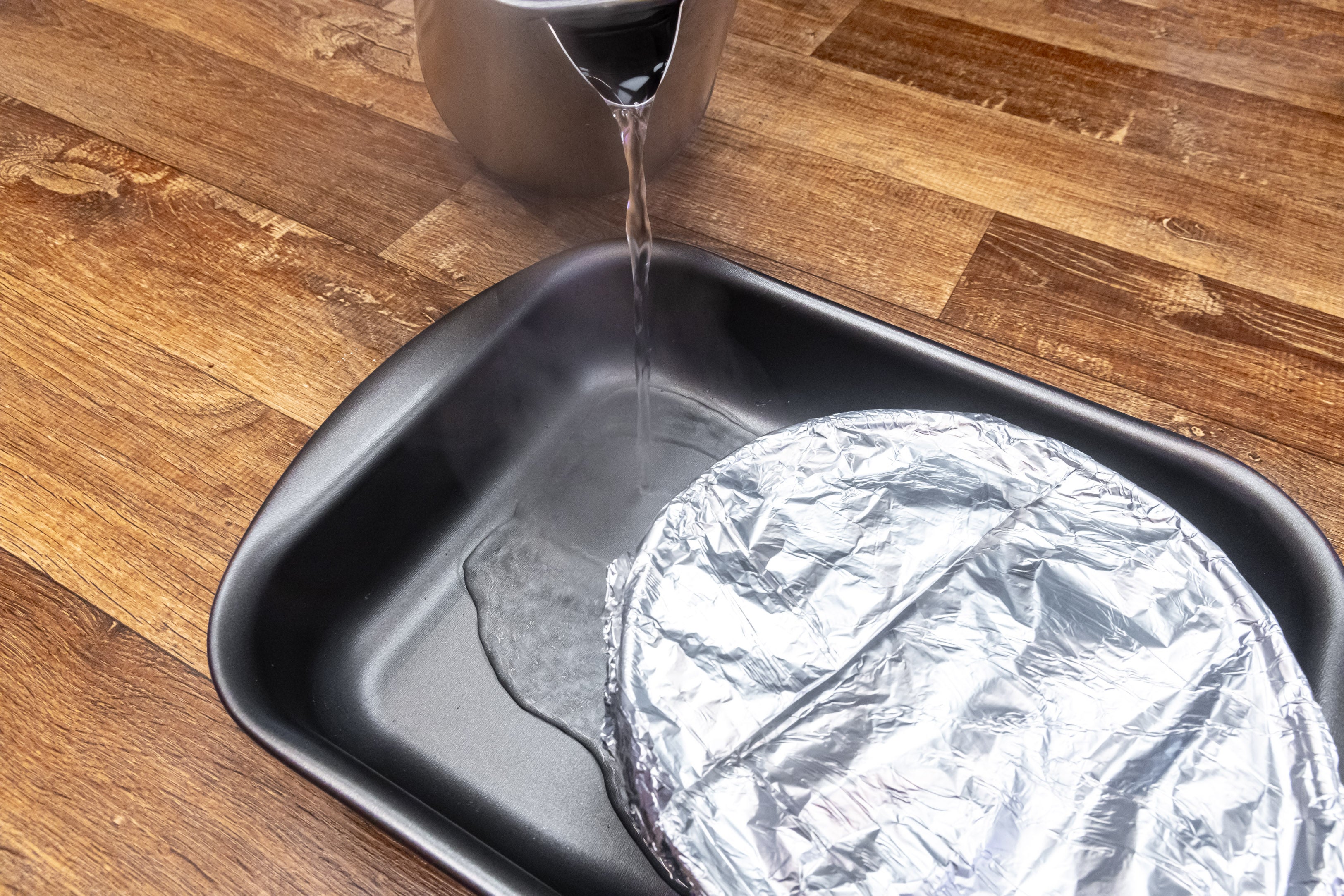 Foto aproximada de uma forma coberta com papel alumínio, dentro de outra forma onde está sendo despejada água quente