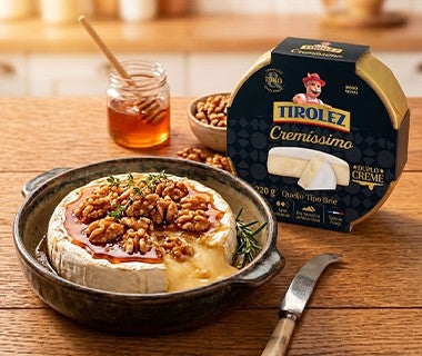 Queijo brie assado com mel e nozes em prato, ao lado de embalagem Tirolez Cremíssimo sobre mesa de madeira	