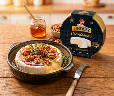 Queijo brie assado com mel e nozes em prato, ao lado de embalagem Tirolez Cremíssimo sobre mesa de madeira	