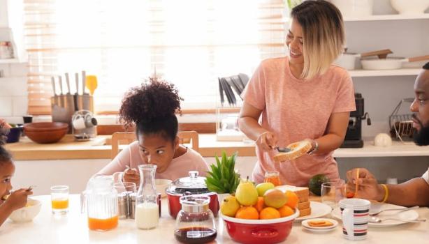 Dicas de café da manhã: Duas crianças negras à esquerda, dois adultos negros à direita, com frutas, leite, pão e café na mesa