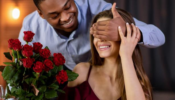 Jantar para o Dia dos Namorados: Homem negro segurando um buquê de flores e com a mão nos olhos de uma mulher branca 