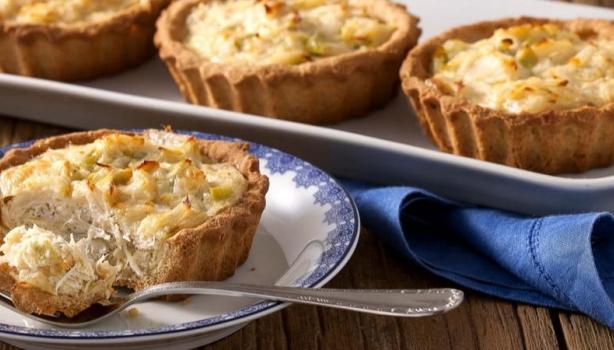 Quiche de alho-poró sendo cortado sobre um prato com um garfo ao lado. Ao fundo, mais 3 quiches inteiros
