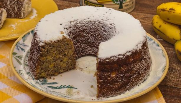 Bolo de banana com um pedaço cortado, à direita 5 bananas e, à esquerda os pedaço do bolo que foram cortados sob um prato