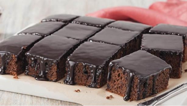 Prato com um bolo de chocolate cortado em pedaços quadrados e uma faca, ao fundo um pano vermelho e um potinho com chocolate 