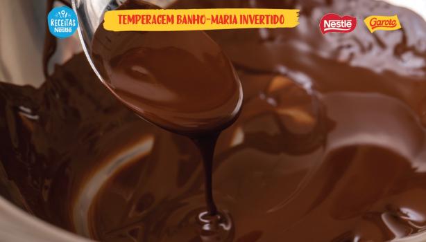 Temperagem em Banho-Maria Invertido Cena bastante aproximada de um recipiente com chocolate derretido escorrendo de uma colher com o título acima