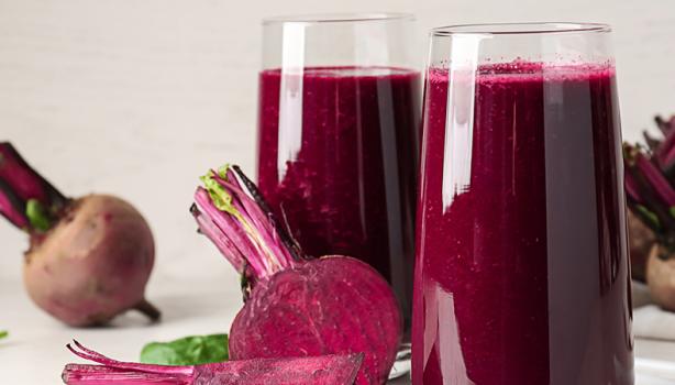 Copos com suco de beterraba, rodeados por beterrabas frescas cortadas e folhas verdes sobre superfície clara.
