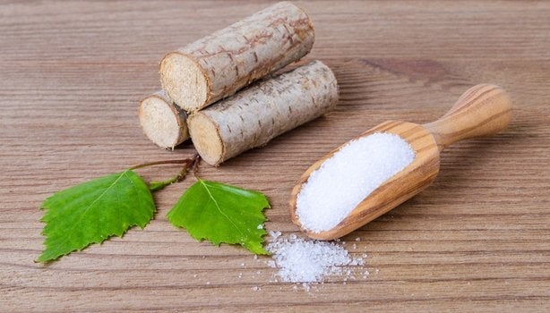 Em uma mesa, há três madeiras empilhadas com duas folhas verdes à frente. Ao lado, temos o xilitol, substituto do açúcar.  