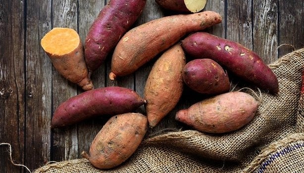 Batatas-doces inteiras e cortadas sobre superfície de madeira rústica com saco de estopa ao lado.