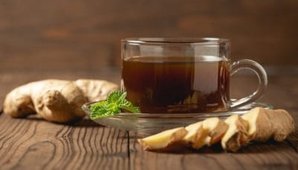 Gengibres inteiros e fatiados ao lado de uma xícara de chá de gengibre. Todos os itens estão em cima de uma mesa de madeira. 