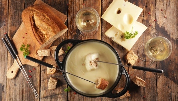 Imagem de uma mesa de madeira com uma panela de fondue e acompanhamentos como pães, queijos e vinho.