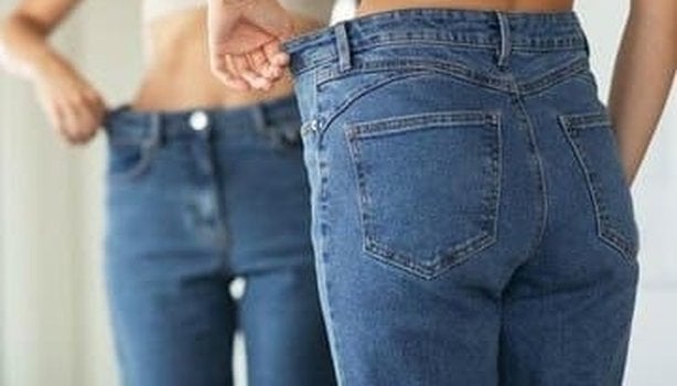 Mulher se olhando no espelho segurando a sobra da calça jeans na lateral. A imagem representa porque não consigo emagrecer.