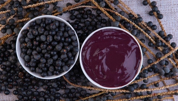 Tigela com polpa de açaí e tigela com frutas de açaí inteiras, sobre ramos com frutos dispostos em superfície clara.