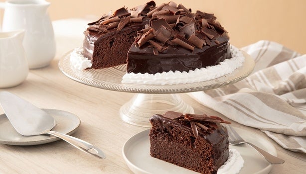 Imagem de um bolo de chocolate com raspas de chocolate sobre uma boleira e uma fatia em um prato.