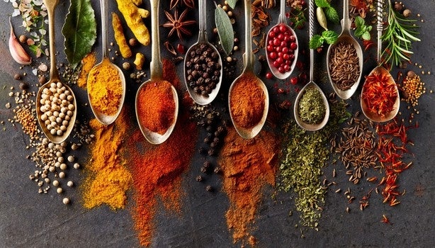 Colheres com diferentes temperos e especiarias coloridas espalhadas sobre superfície escura.