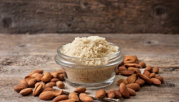 Pote de vidro com farinha de amêndoas, cercado por amêndoas cruas sobre superfície de madeira rústica.