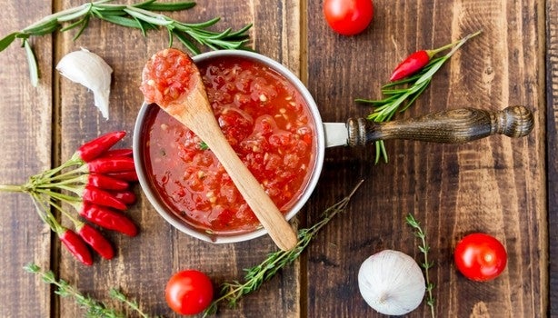 Imagem de uma panela com molho de tomate com uma colher de pau sobre uma superfície de madeira com tomates.