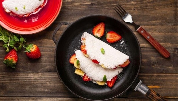 Duas tapiocas recheadas com frutas em uma frigideira sobre uma mesa de madeira. Ao lado, um prato com outra tapioca.