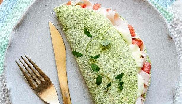 Imagem de uma tapioca verde de espinafre recheada com queijo e tomate sobre um prato cinza.