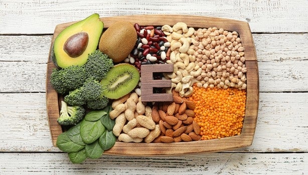 Alimentos ricos em vitamina E em bandeja de madeira: abacate, kiwi, brócolis, espinafre, castanhas e leguminosas.