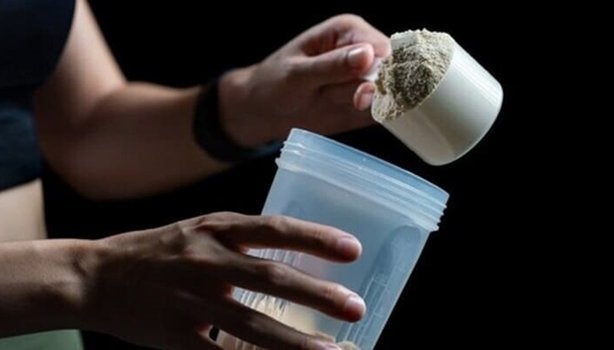 Mulher segurando um copo de plástico com Whey Protein, e com a outra mão segura um medidor cheio da mesma proteína.