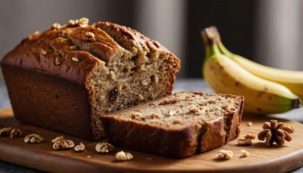 Bolo de banana com nozes fatiado em tábua de madeira, decorado com pedaços de noz e bananas ao fundo.