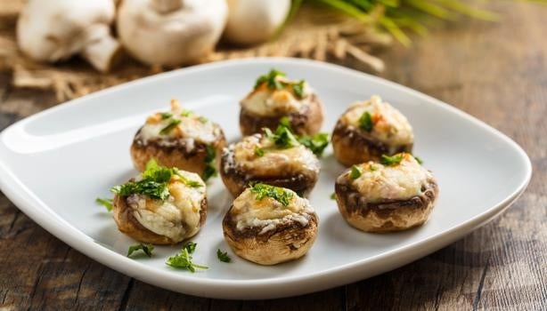 Cogumelos recheados com queijo e ervas frescas servidos em prato branco sobre mesa rústica de madeira.