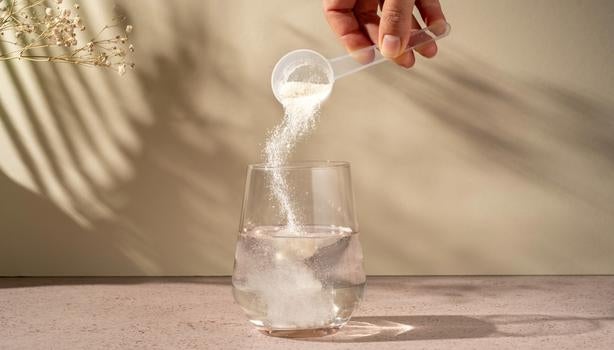 Mão despejando pó de colágeno de uma colher medidora em um copo de água transparente sobre a mesa.