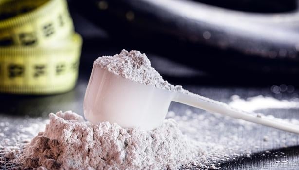 Colher medidora com creatina em pó branca sobre a mesa, ao lado de uma fita métrica amarela desfocada.