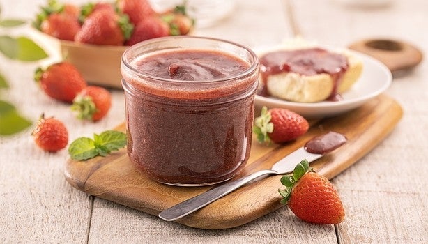 Pote de geleia de morango sobre tábua de madeira, com morangos frescos e pão com geleia ao fundo.