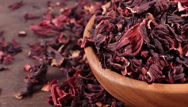 Pétalas secas de hibisco em uma tigela de madeira, com flores espalhadas ao redor sobre superfície de madeira.