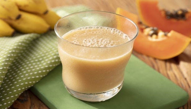 Vitamina cremosa servida em copo transparente, com bananas e fatias de mamão ao fundo sobre mesa de madeira.