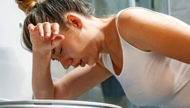 Em um banheiro, uma mulher está apoiada no vaso sanitário, de olhos fechados, mostrando um sofrimento com virose.