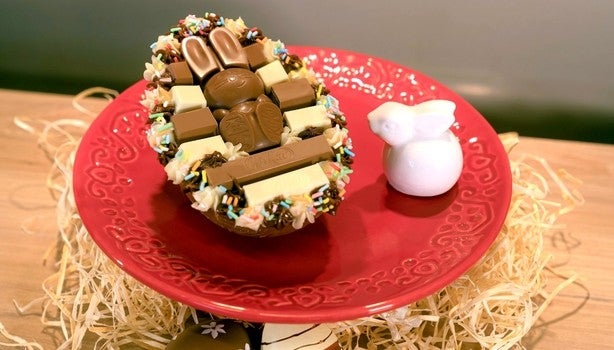 Ovo de Páscoa recheado e decorado com chocolates KitKat e confeitos coloridos, servido em prato vermelho com coelho branco.