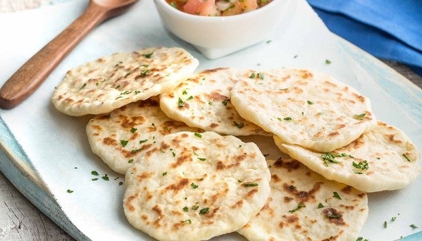 Imagem de pães achatados dourados servidos em prato branco, decorados com salsa e acompanhados de molho em tigela.